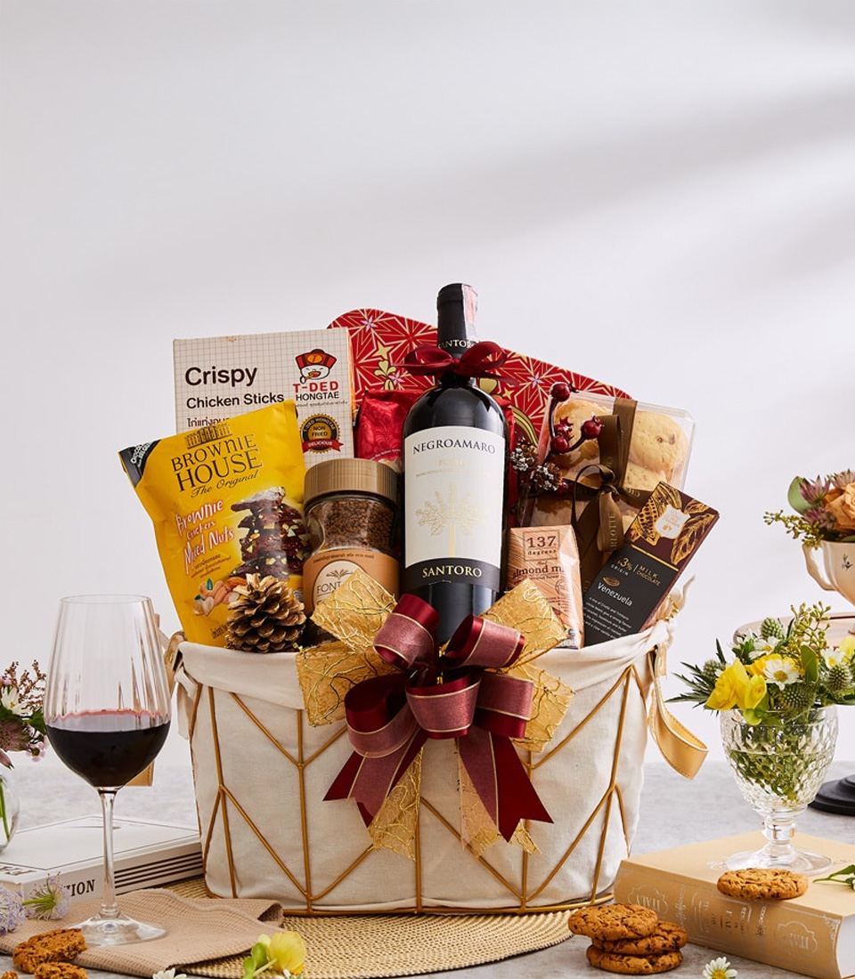 A lavish gift basket from Basketeer graces a pristine white table, exuding elegance and opulence. Nestled within are gourmet delights, including a bottle of exquisite red wine, savory chicken sticks, delectable breadsticks, and indulgent cookies. Each item is carefully arranged and adorned with a sumptuous red and gold ribbon featuring the Basketeer logo, adding a touch of sophistication. The scene is complemented by a glass of rich red wine alongside delicate blooms and an inviting plateful of cookies, creating an atmosphere of pure luxury and warmth.
