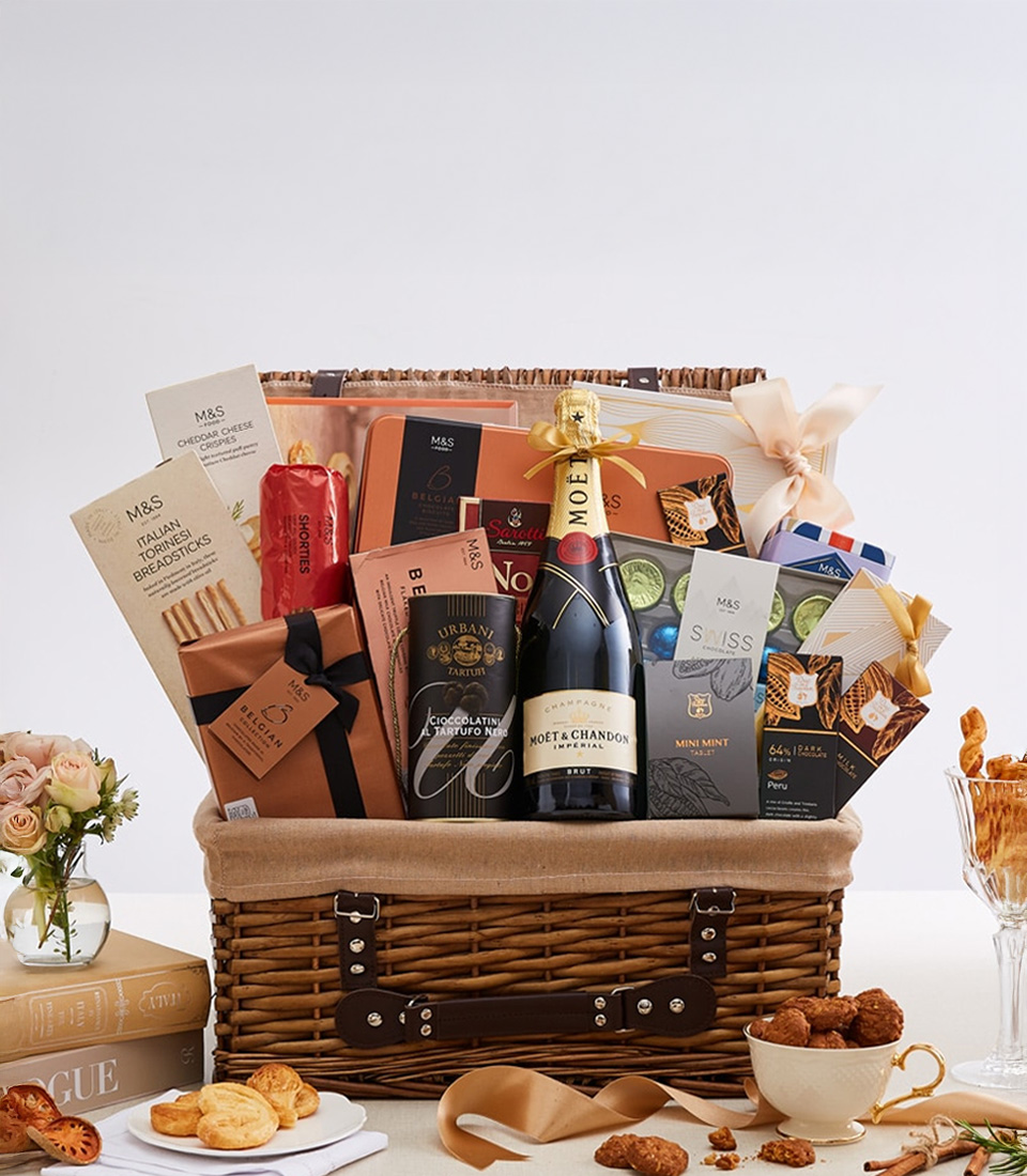 An opulent Basketeer gourmet gift basket overflowing with decadent treasures: an exquisite bottle of Moët & Chandon champagne, a delightful selection of artisanal chocolates, buttery biscuits, fine teas, and gourmet delicacies, elegantly nestled in a generous wicker hamper. The scene is beautifully set with lush flowers, classic books, and additional tantalizing snacks. A luxurious ribbon adorned with the Basketeer logo adds a touch of refinement to this sumptuous display.