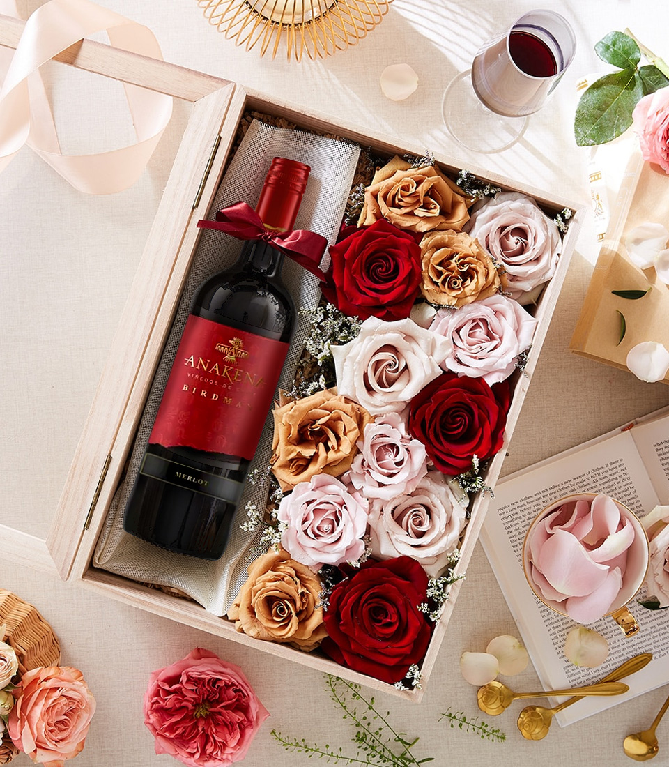 A beautifully arranged wooden gift box featuring a bottle of Anakena Birdman Merlot wine, adorned with a deep red ribbon, surrounded by luxurious preserved roses in shades of red, blush, and champagne. A glass of red wine and decorative floral elements complete the romantic ambiance.