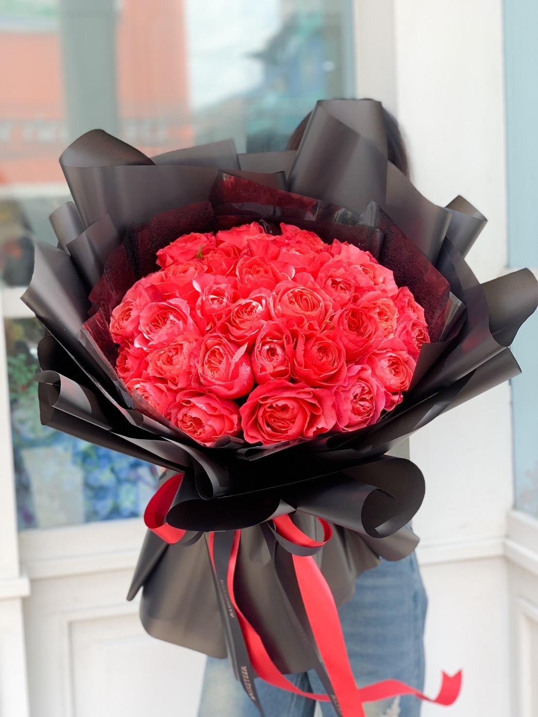 A person is holding a large bouquet of vibrant Love's Red Explorer Roses wrapped in black and translucent plastic with red ribbons. The bouquet is positioned at the center, obscuring the person's face and upper body. The background features a white wall and glass window.