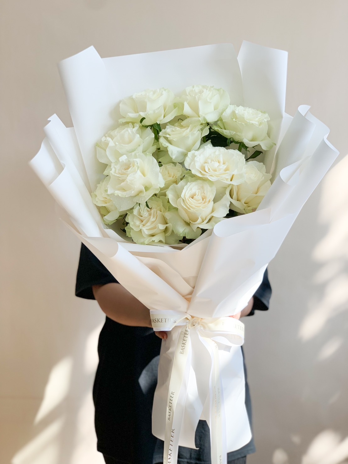 A beautifully arranged bouquet of fresh white roses, elegantly wrapped in white paper with a Basketeer ribbon, held by a person dressed in black.