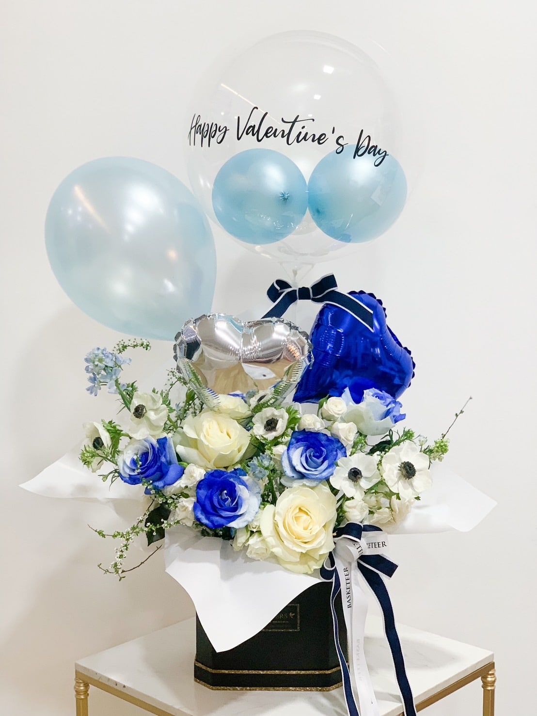 A floral arrangement in a black box featuring white and blue roses, anemones, and greenery. Four balloons are attached: one large clear balloon with 