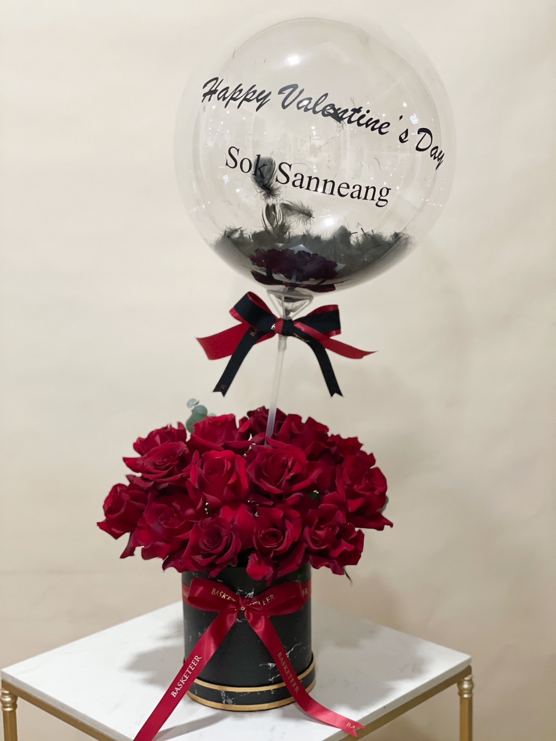 A luxurious romantic gift featuring a box of fresh red roses with a personalized clear balloon, beautifully decorated with black feathers and ribbons.