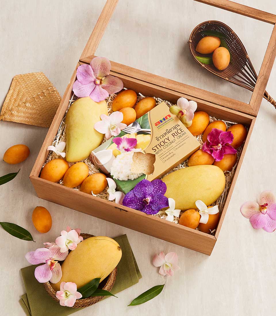A Songkran Blissful Wood Box with a transparent lid contains ripe mangoes, small orange fruits, a package of Thai coconut sticky rice, and orchid flowers for decoration. Nearby, a woven mat and a dish with more orange fruits rest on a green napkin.
