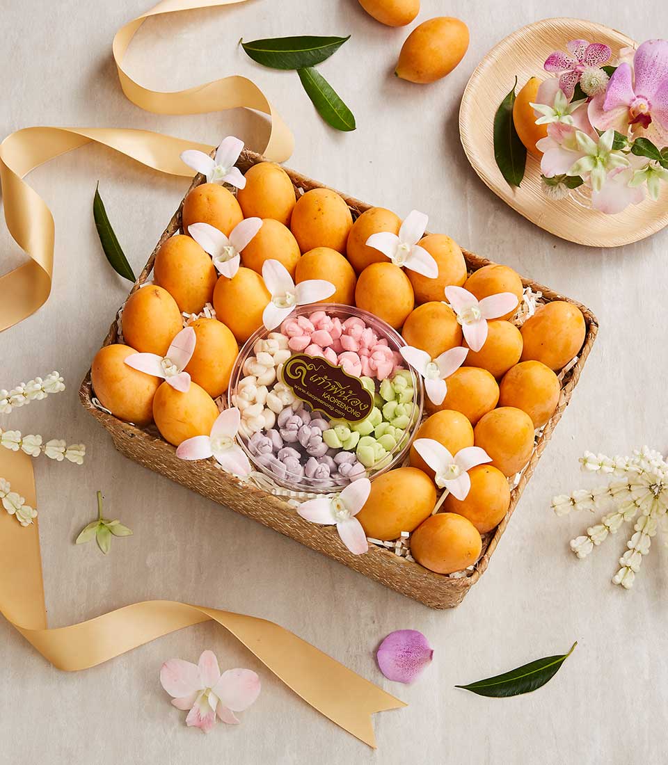 A woven Thai Songkran Gift Basket filled with ripe yellow mangoes and a container of assorted Thai desserts sits on a light-colored table. The basket is adorned with white flower decorations and surrounded by golden ribbons, green leaves, and pink orchids.