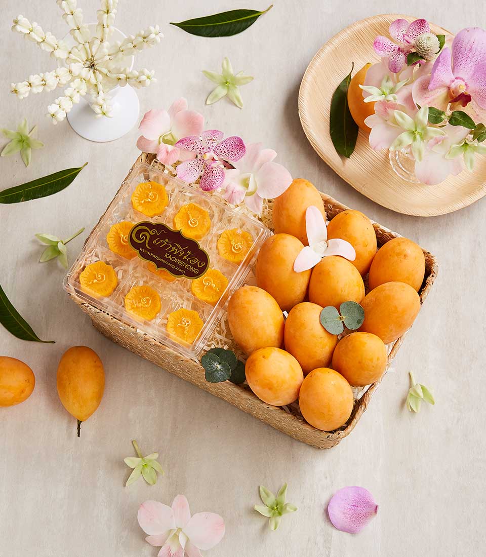 A Thai dessert basket holds several ripe marian plums next to a clear box of delicately arranged yellow flowers. Surrounding the basket are scattered green leaves, white jasmine blooms in a vase, and light pink orchids on a plate and as decoration.