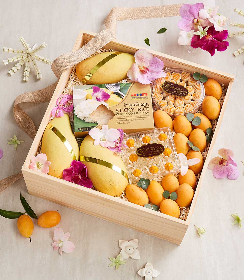 A Thai New Year Blessings Gift: A wooden gift box containing ripe mangoes, sticky rice packs, and other snacks, garnished with pink and white flowers. The box is adorned with a beige ribbon, while small green leaves and white flower petals are scattered around for a festive touch.