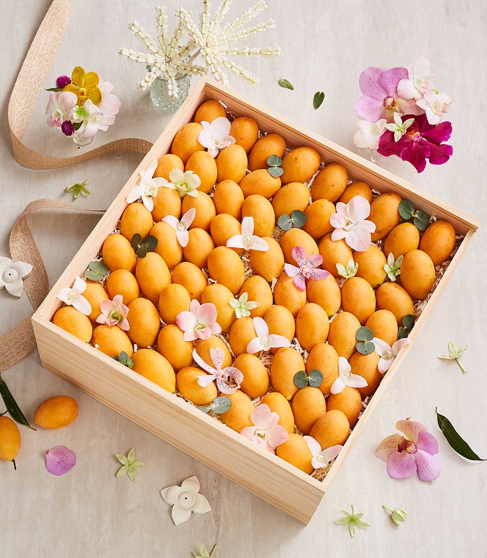A wooden box filled with ripe, sweet Marian plum fruit sits on a light surface. The plums are arranged in neat rows and adorned with delicate pink and white flowers. Surrounding the gift box set are scattered flowers and green leaves, enhancing its decorative presentation.