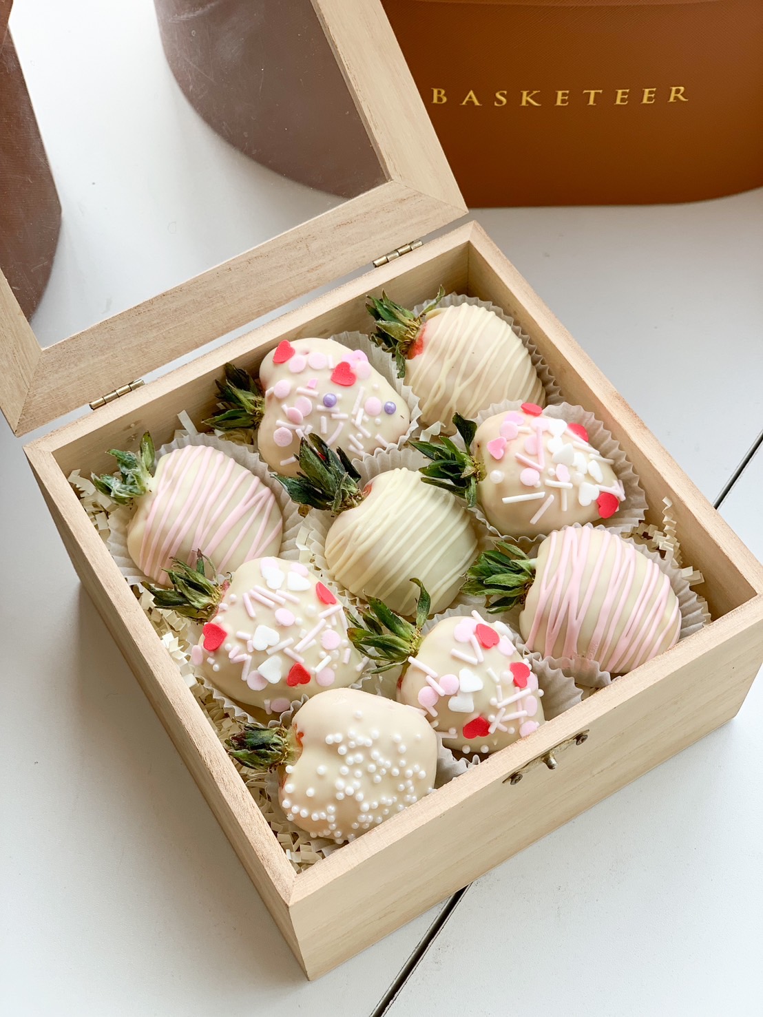 A wooden gift box filled with white chocolate-covered strawberries, decorated with sprinkles, heart-shaped toppings, and drizzles.