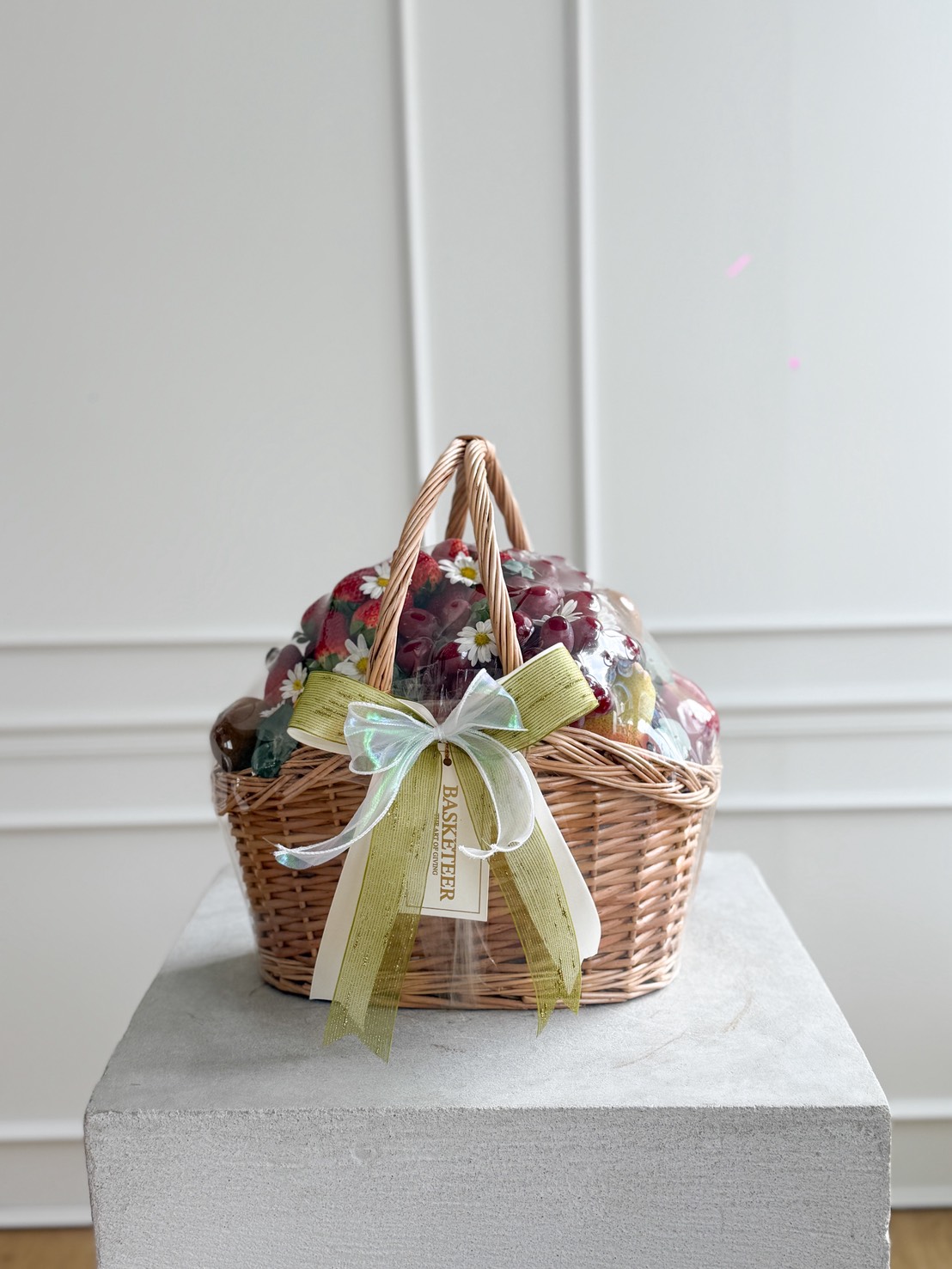 A wicker hamper filled with mixed luxury fresh fruit, wrapped in clear plastic and adorned with a green and white bow and a 