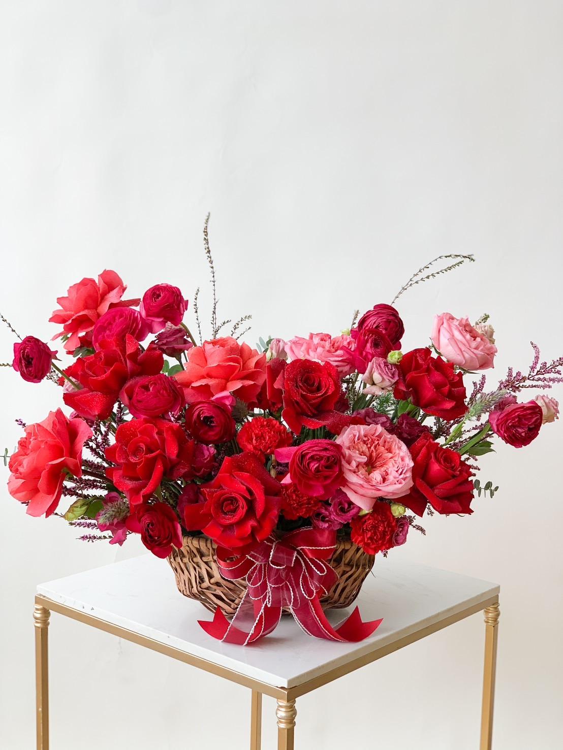A wicker basket overflowing with a variety of vibrant red and pink flowers, including roses and carnations, accented with green foliage. The arrangement is adorned with a red ribbon bow and is placed atop a small white table with light-colored legs.