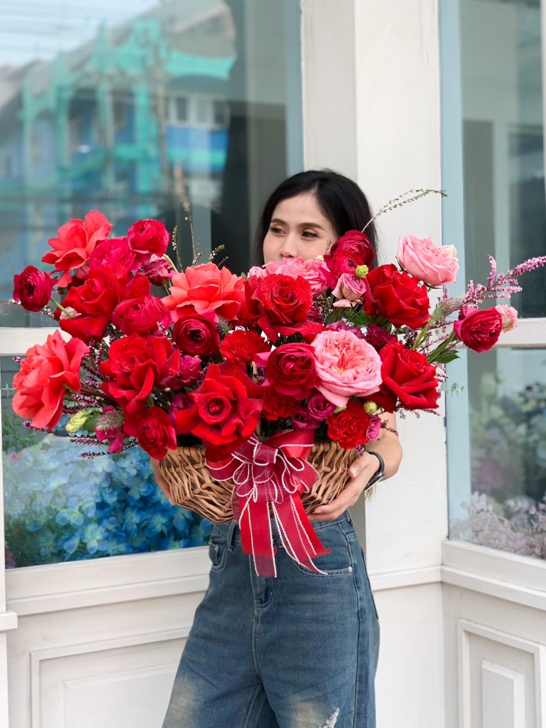 A woman stands outdoors holding a large, colorful bouquet of red, pink, and coral roses in a brown basket adorned with a red ribbon. She is wearing a dark top and blue jeans, and the background shows a storefront with various flowers.