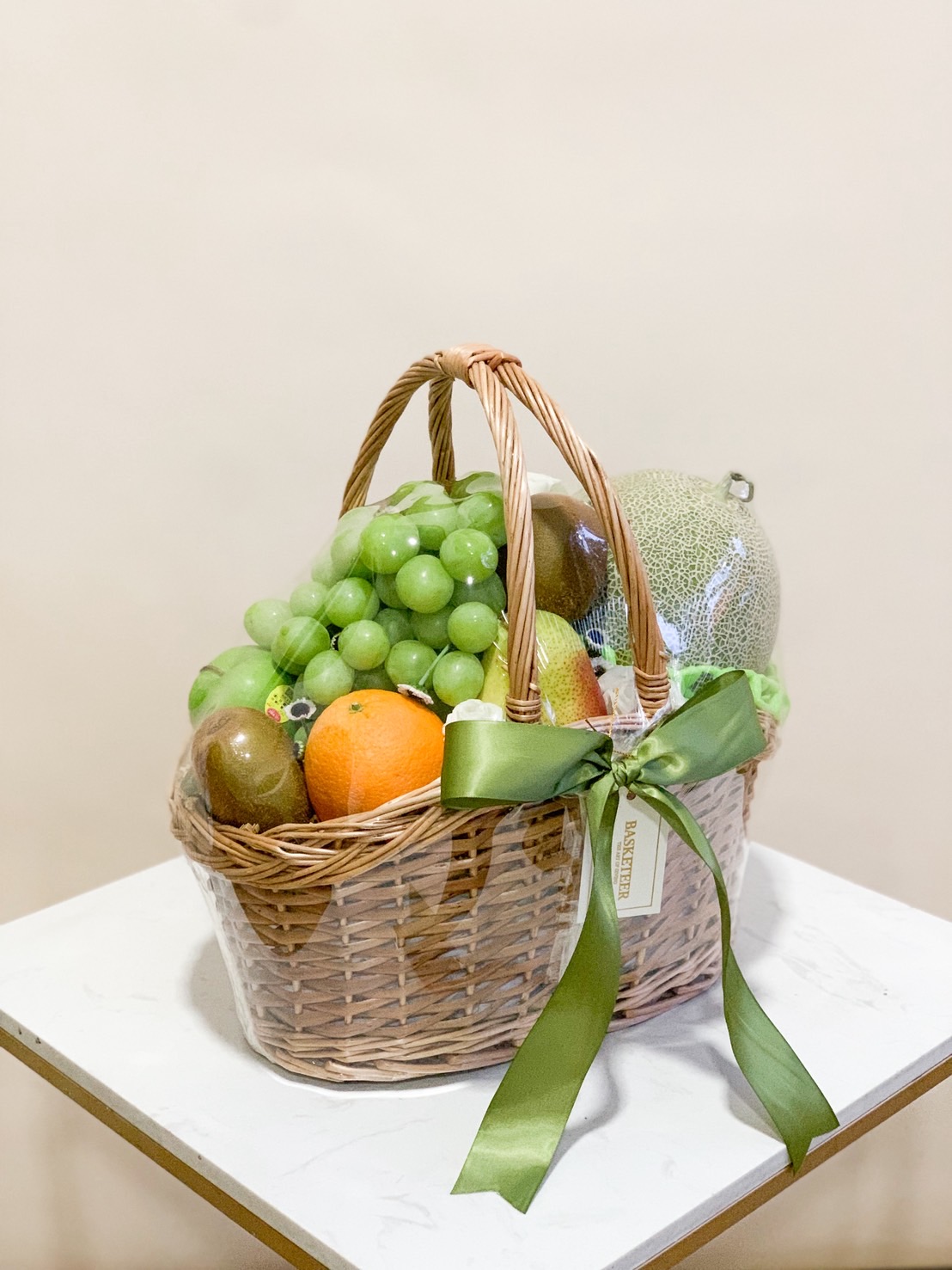 Luxury premium fruit basket with green grapes, orange, kiwi, pear, and melon, decorated with green satin ribbon.