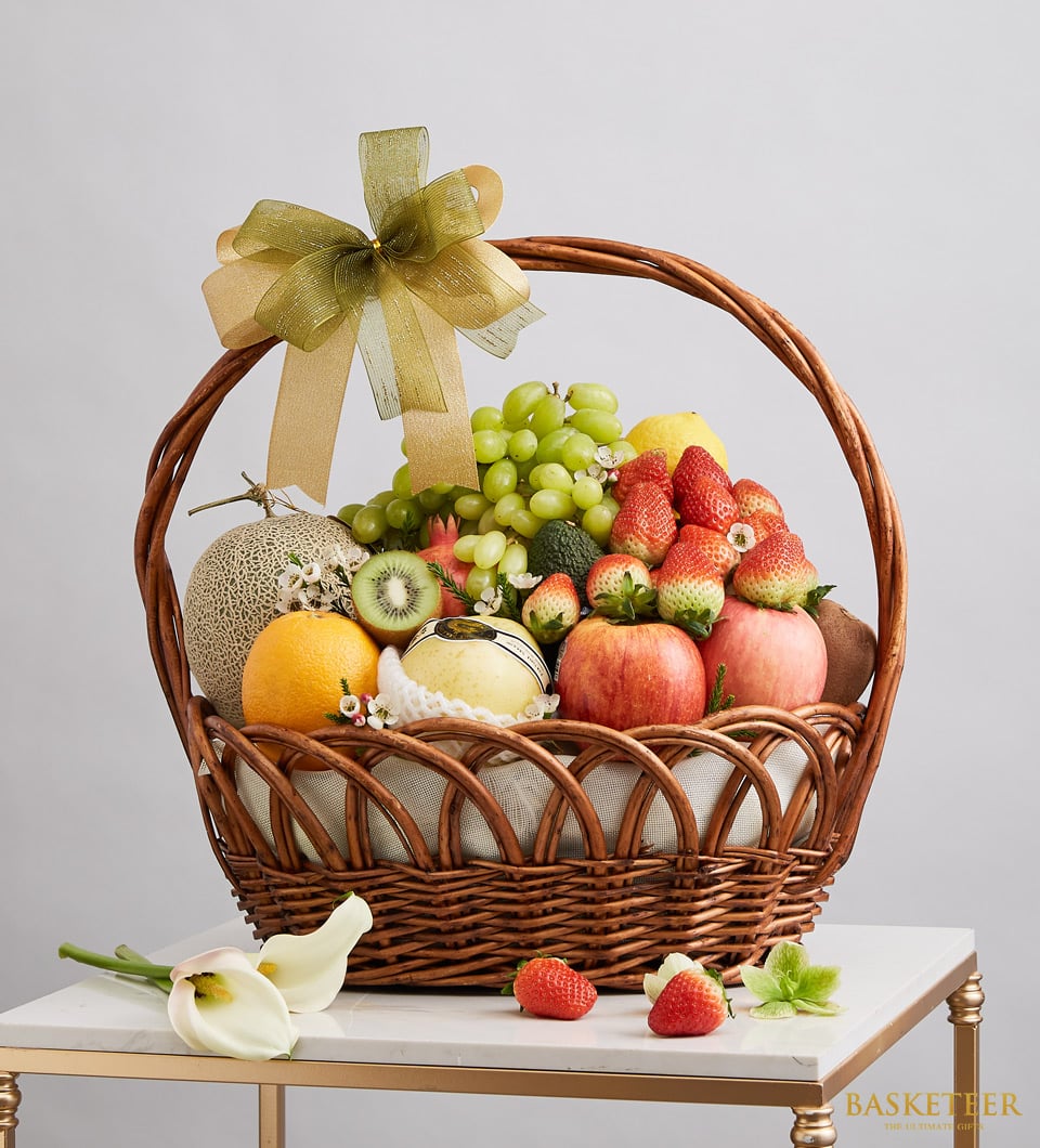 An exquisite woven basket overflowing with a sumptuous assortment of premium fruits: vibrant green grapes, succulent strawberries, crisp apples, a perfectly ripe melon, a luscious pear, and a zesty orange. The opulent Regal Fruit Gift Basket from Basketeer is elegantly adorned with a luxurious gold ribbon emblazoned with the Basketeer logo. The basket is tastefully situated amidst delicate white flowers and artfully arranged scattered fruit on an immaculate white surface.