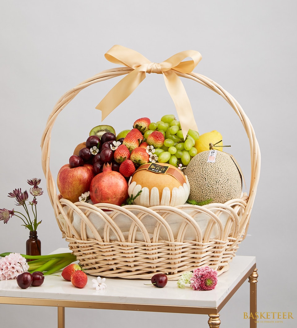 A luxurious and enchanting Basketeer fresh fruits basket, brimming with a succulent assortment of crisp apples, juicy grapes, luscious strawberries, an exotic mango, and fragrant melons. This elegant arrangement is topped off with a sophisticated beige satin bow bearing the Basketeer logo. The enticing display rests gracefully on a pristine white table beside a charming bouquet of vibrant purple flowers in an exquisite glass vase.