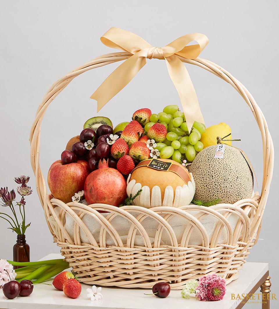 An exquisite wicker gift basket by Basketeer, elegantly adorned with a luxurious yellow ribbon. This opulent arrangement overflows with an array of fresh, succulent fruits – crisp apples, luscious strawberries, plump grapes, and ripe cantaloupe. A melon wrapped in a delicate ribbon adds an extra touch of sophistication. The scene is completed with scattered blooms that enhance the allure, all set against a simple yet refined backdrop that emphasizes the basket's grandeur and charm.