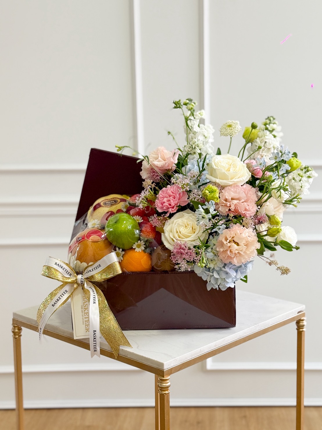 A luxurious gift box filled with assorted fresh fruits including oranges, apples, grapes, and a large pear, paired with an elegant floral arrangement of white roses, pink carnations, peach blooms, blue hydrangeas, and delicate filler flowers. The box is decorated with a gold and white ribbon and displayed on a marble-top table against a white paneled background.