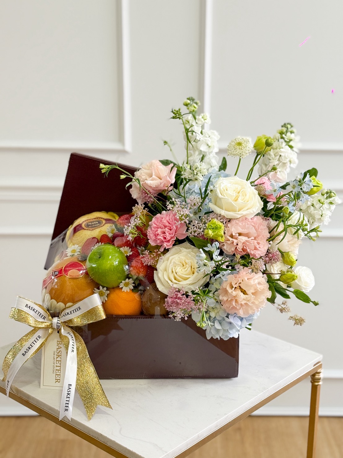 A luxury gift box filled with assorted fresh fruits such as green apples, oranges, grapes, and a premium pear, paired with a pastel floral arrangement featuring white roses, pink carnations, peach-toned blooms, blue hydrangeas, and delicate filler flowers. The box is decorated with a gold and white ribbon and displayed on a marble-top table against a white paneled background.