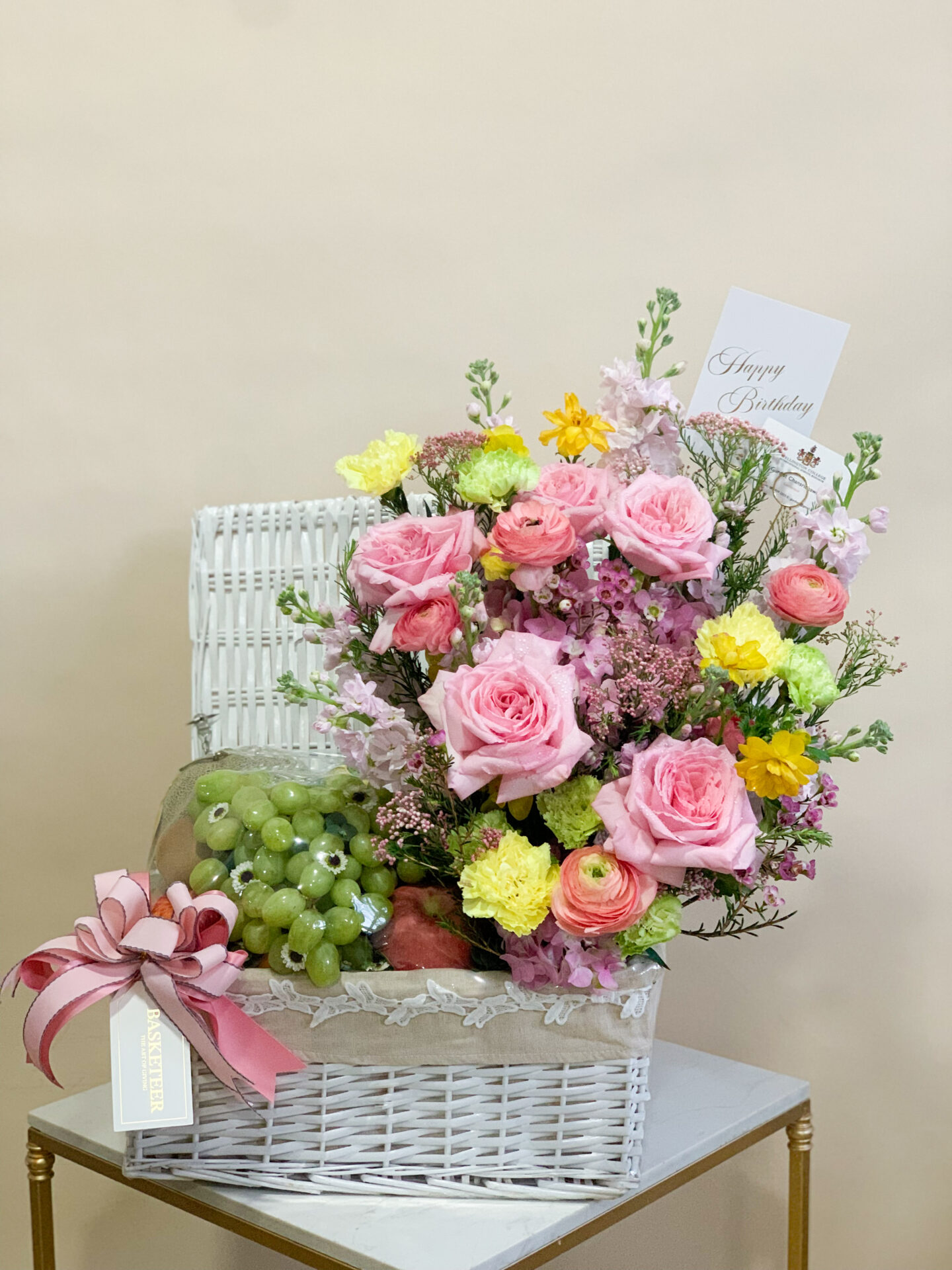 A sophisticated woman in an elegant white turtleneck sweater and chic pink skirt cradles an opulent floral fruit basket from Basketeer. Adorned with a luxurious pink ribbon showcasing the Basketeer logo, this exquisite arrangement brims with vibrant roses and tulips mingling harmoniously with fresh, succulent fruits like grapes and oranges. The scene exudes luxury and warmth, making it irresistibly inviting.