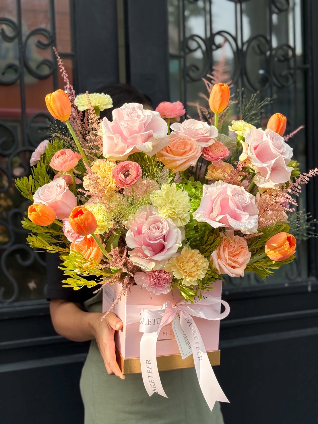 Pastel floral gift box with pink roses, orange tulips, peach carnations, and yellow ranunculus in pink square box