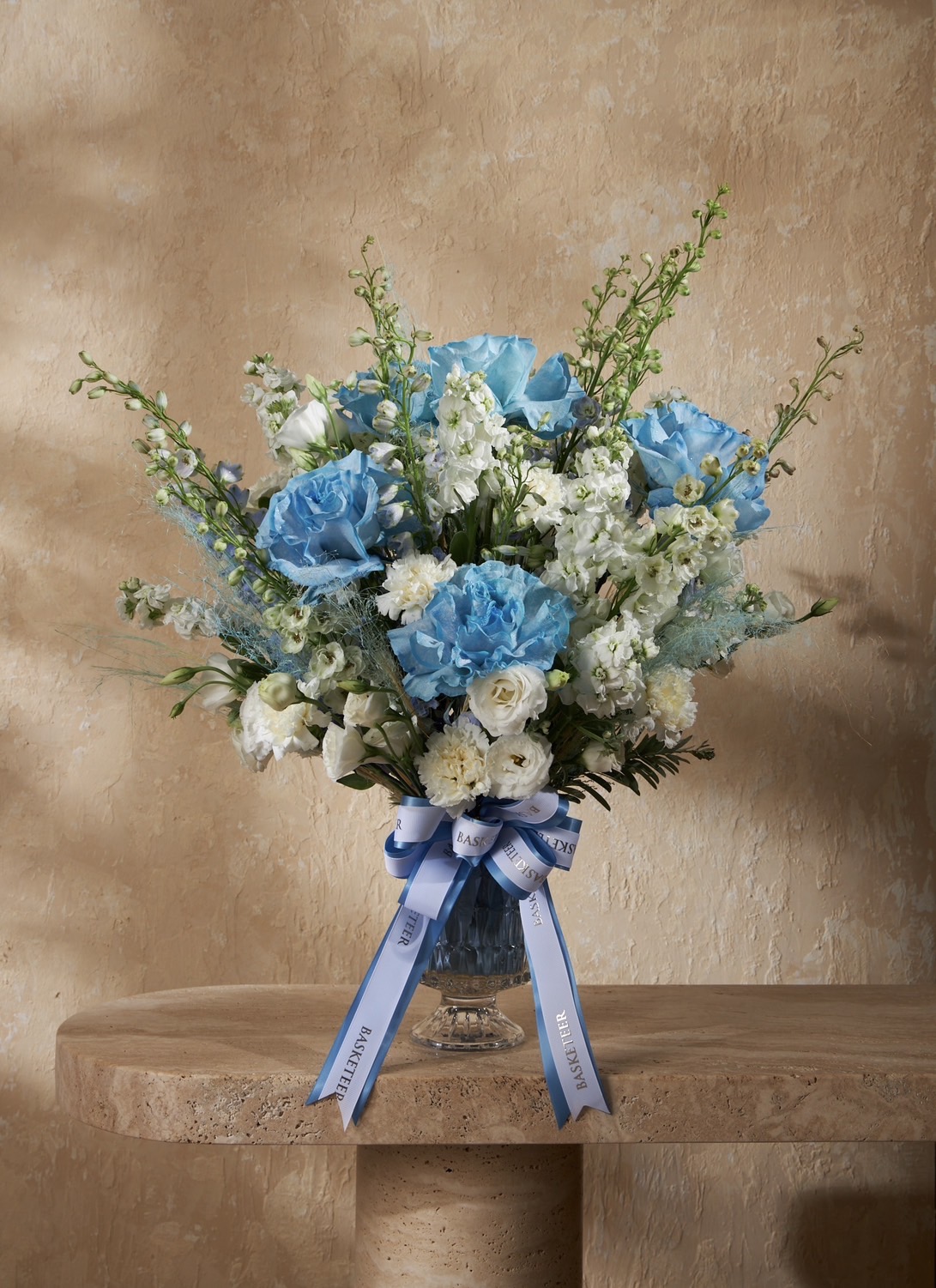 Elegant blue and white roses in a glass vase with soft greenery, styled with satin ribbons on a luxury stone table.