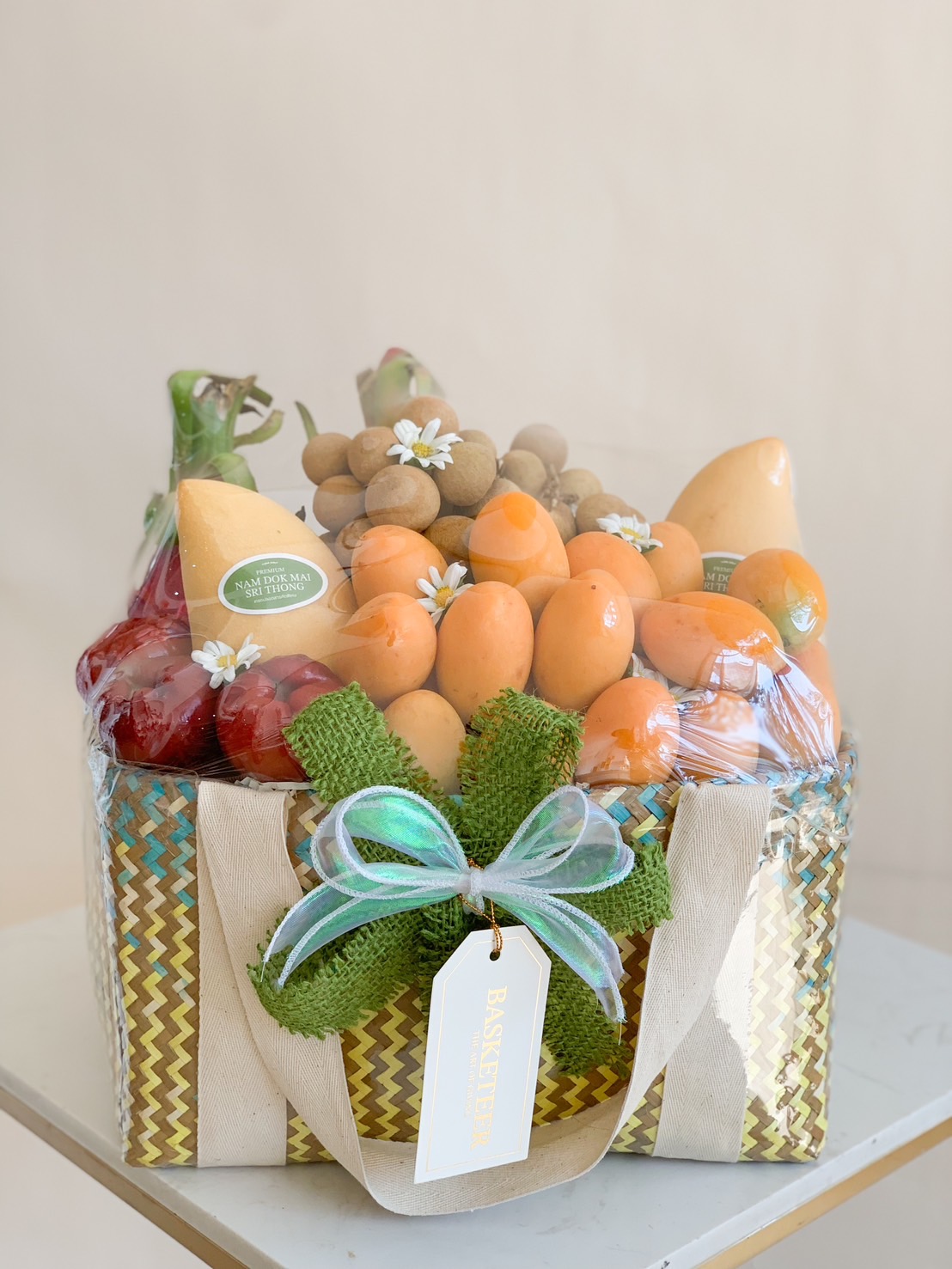 A beautifully arranged tropical fruit basket, featuring fresh mayongchid (Marian plums), longan, mangoes, grapes, and bananas, wrapped in an elegant woven basket with a green ribbon and floral accents—perfect for gifts and celebrations.