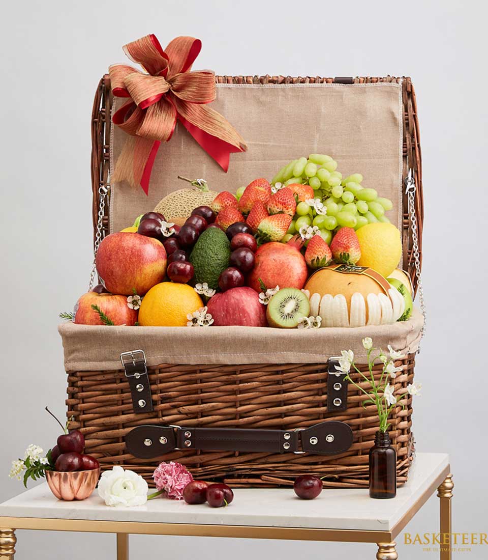 Brown rattan gift basket with a lid. Filled with various fruits such as red cherries. red strawberry Green grapes and more, decorated with red and sparkling pale gold ribbon bows.