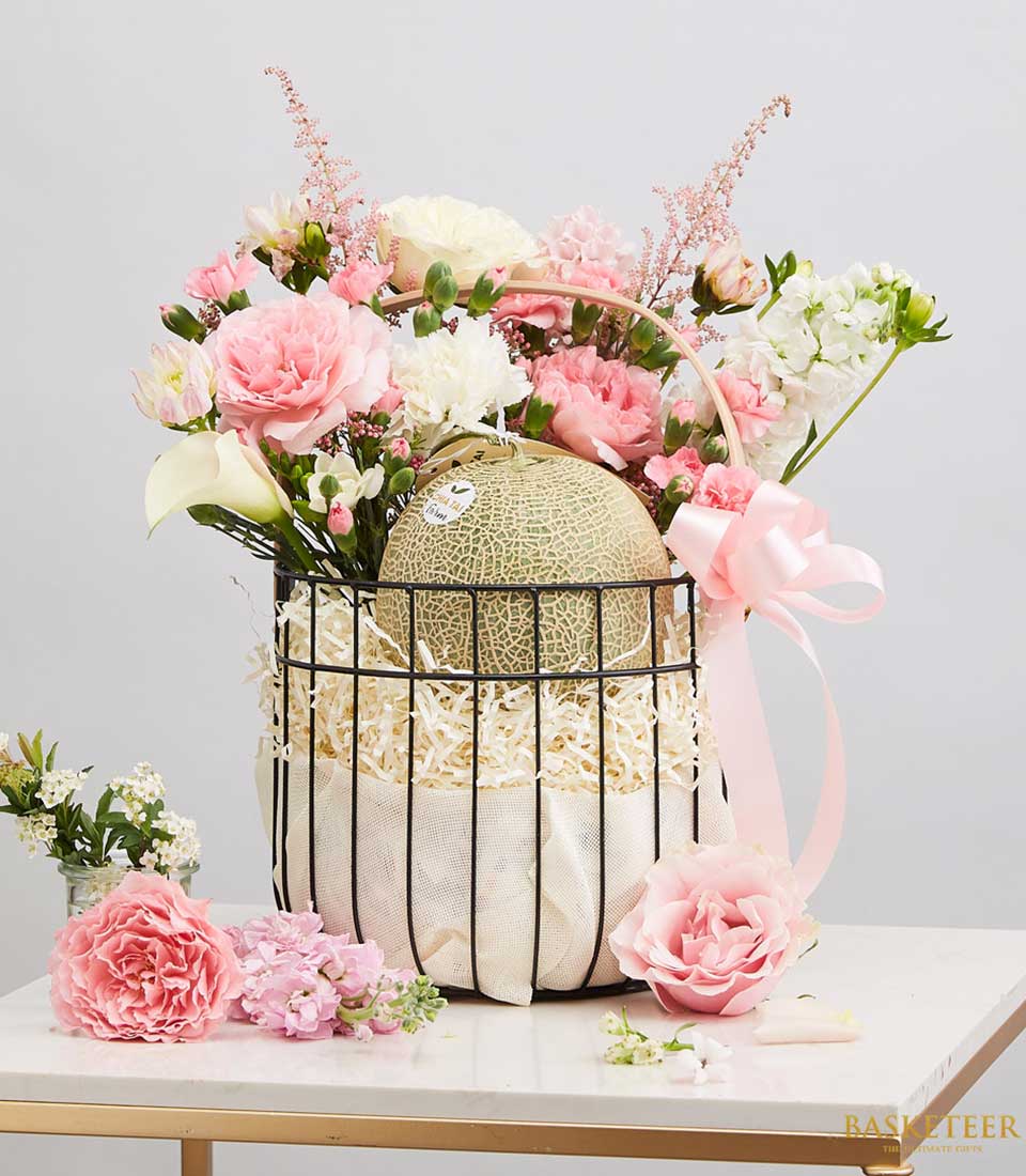 Black Steel Frame Gift Basket Filled with fresh fruits green melons With light pink roses and white flowers Decorated with a light pink ribbon bow.