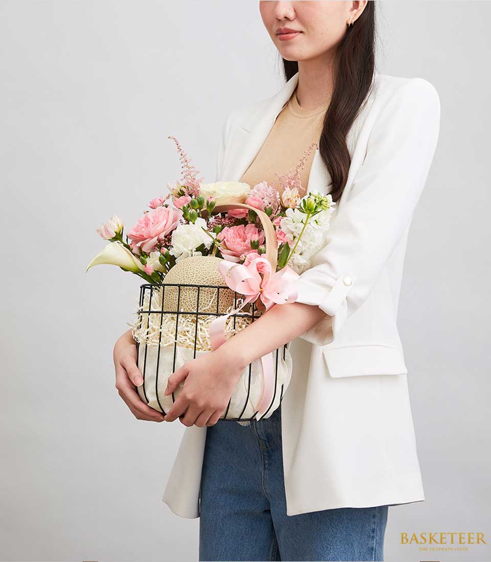 Black Steel Frame Gift Basket Filled with fresh fruits green melons With light pink roses and white flowers Decorated with a light pink ribbon bow.