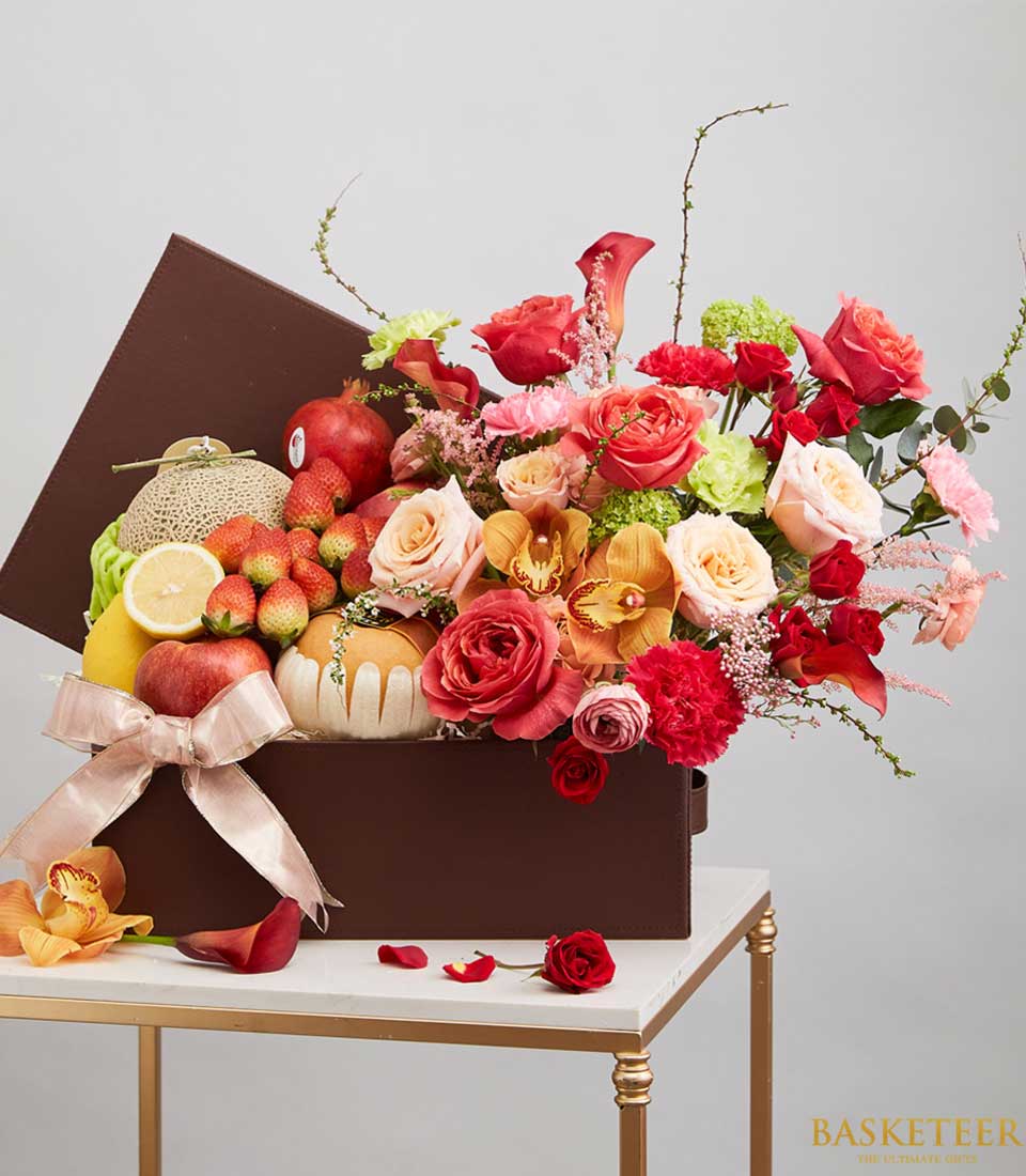 A big dark brown gift box Filled with various types of fresh fruits such as green melons, red pomegranates, and many more, along with red-orange roses. and colorful flowers Decorated with a light brown ribbon bow.