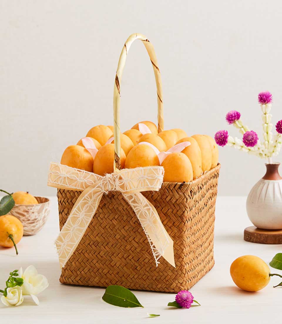 A woven basket filled with ripe yellow mangoes, elegantly presented as a classy fruit gift. The basket, adorned with a light-colored ribbon tied into a bow, sits against a backdrop featuring a small vase of purple pom-pom flowers and scattered green leaves on the white surface.