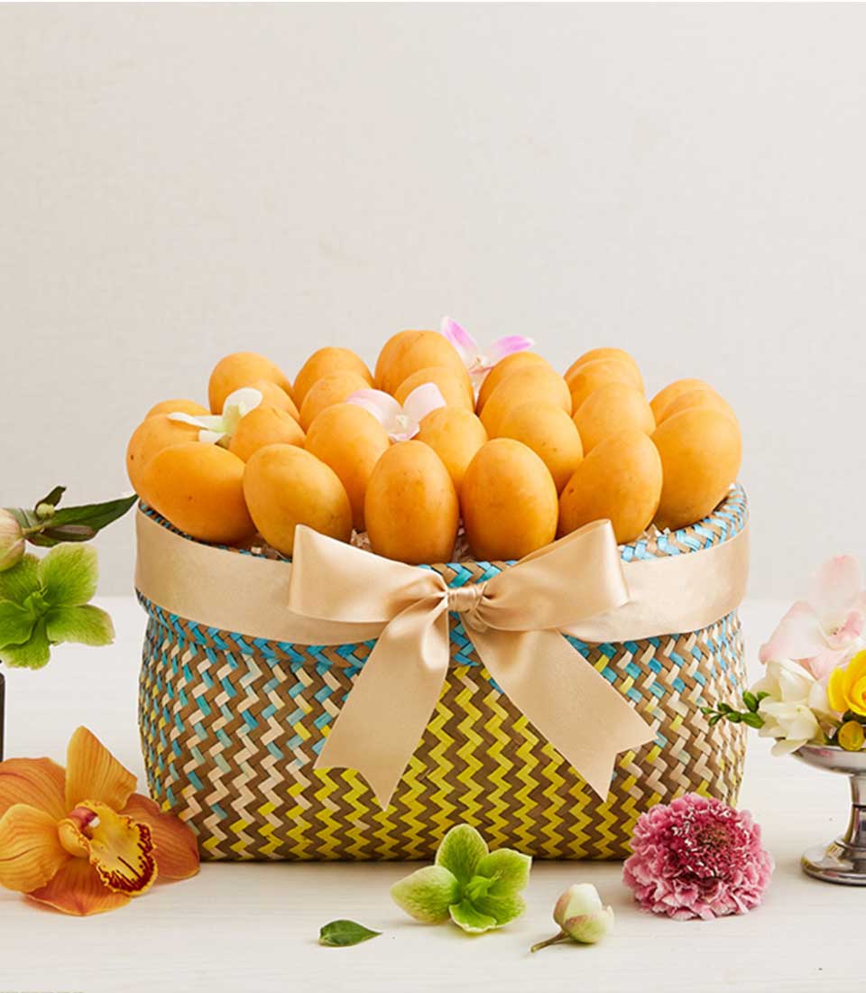 A luxury Songkran Serenade Celebration Basket by Basketeer, overflowing with succulent, golden mangoes and elegantly adorned with a refined beige ribbon bearing the Basketeer logo. This opulent display is surrounded by an enchanting array of vibrant flowers nestled in delicate glass vases, all arranged meticulously on a pristine white surface. The serene backdrop of the light-colored wall enhances the rich, inviting atmosphere of this exquisite scene.