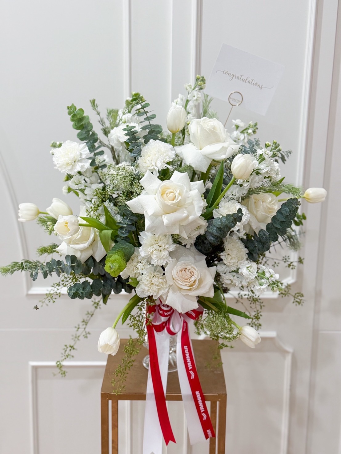 Elegant white floral arrangement featuring roses, tulips, carnations, and eucalyptus in a glass vase with red and white ribbon.