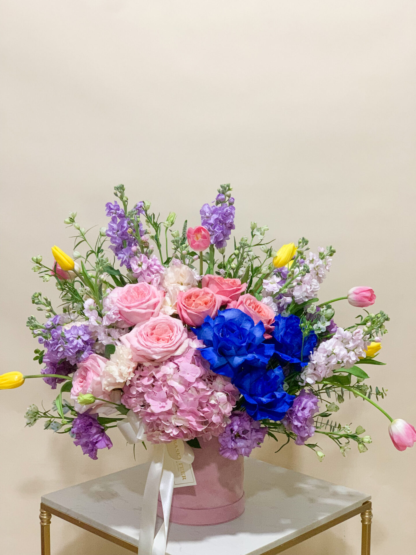 Pink roses, blue hydrangeas, yellow tulips, and lavender blooms arranged in a pink velvet hatbox on a table.