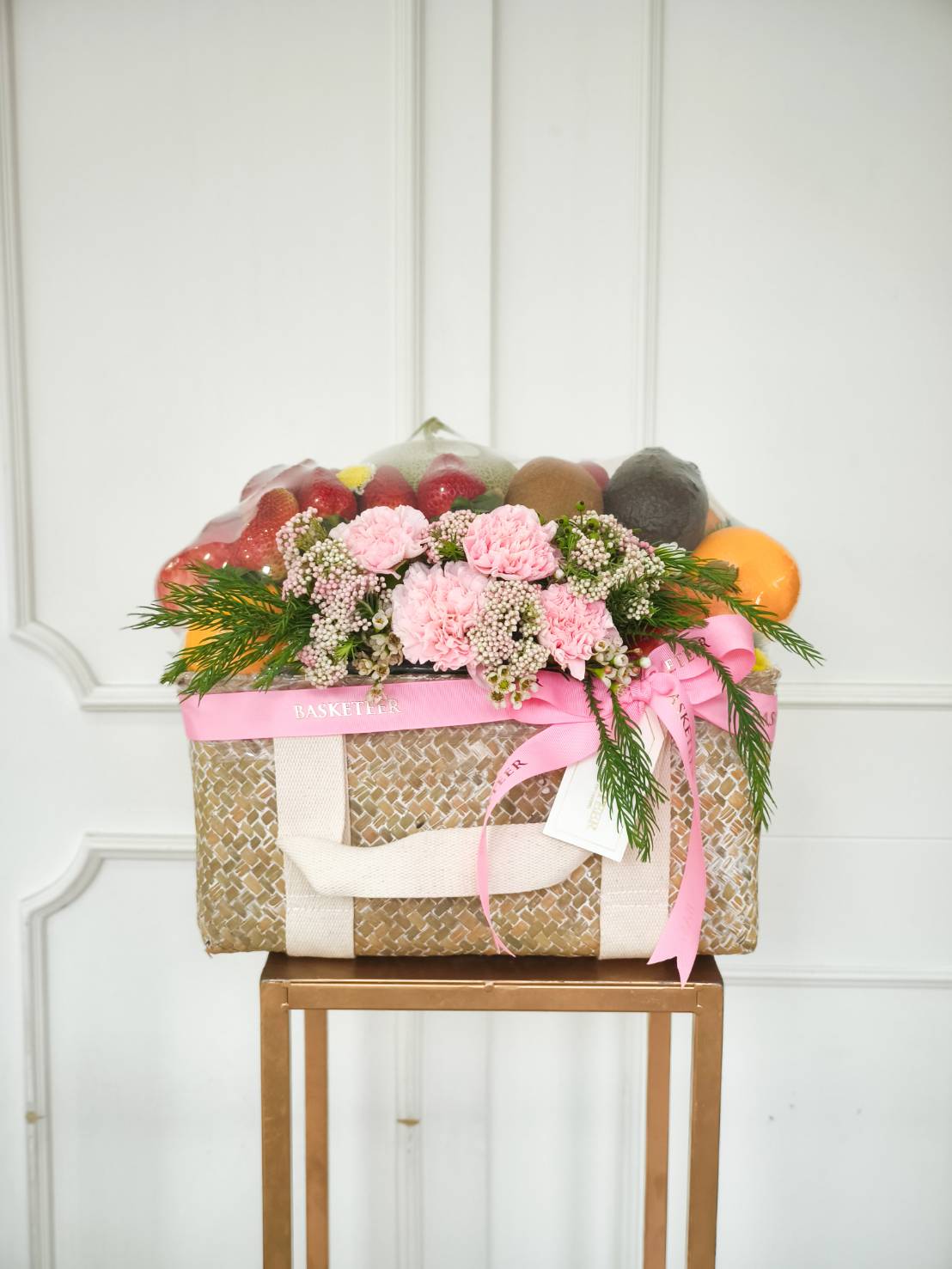 Luxury fruit basket with assorted fresh fruits, decorated with pink carnations, waxflowers, and greenery, tied with a pink satin ribbon.