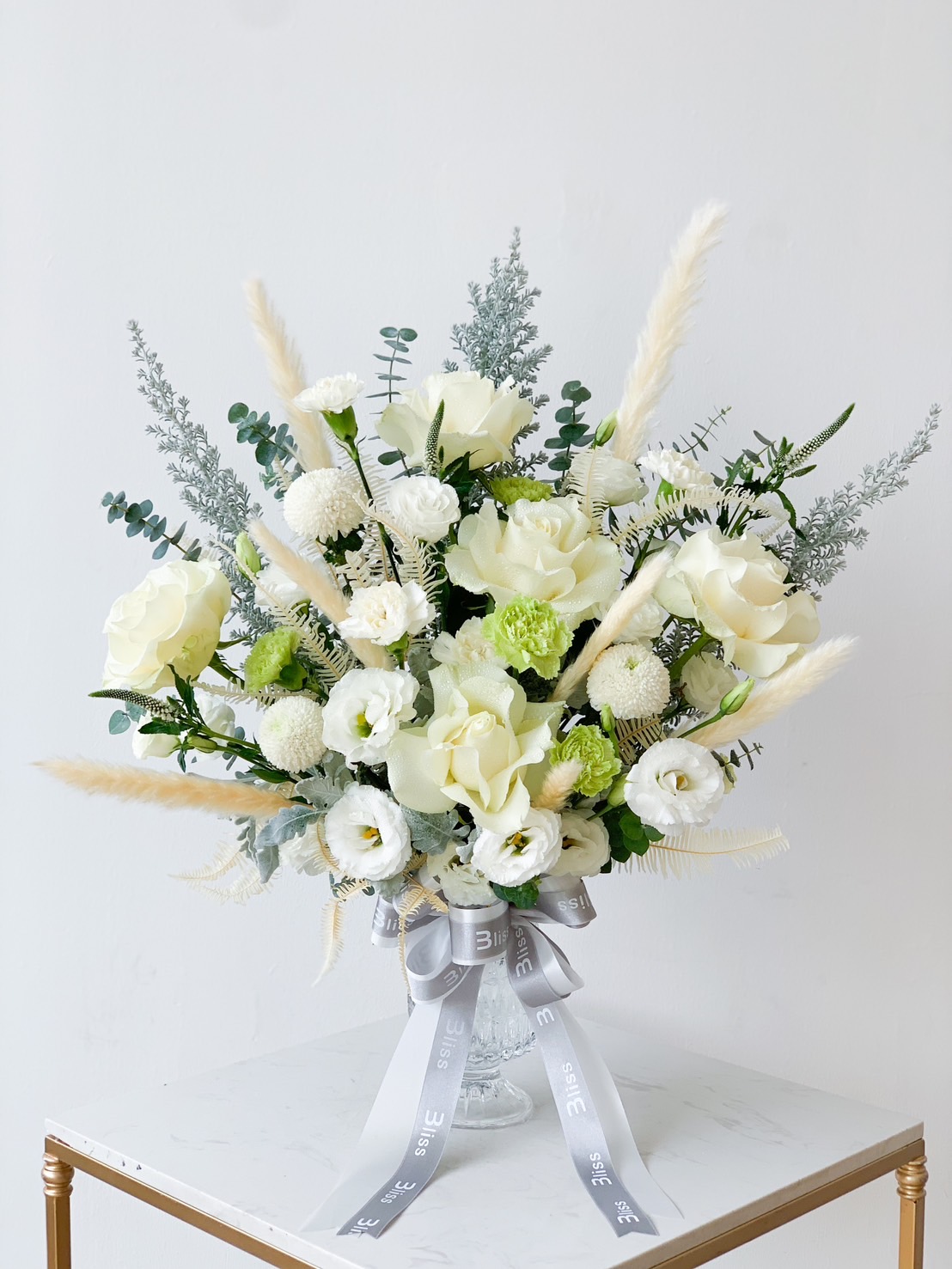 A bouquet of white flowers, including roses and carnations, arranged with green foliage and white pampas grass. The bouquet is wrapped with a silver satin ribbon and displayed in a clear glass vase. It is placed on a white table against a light grey background.