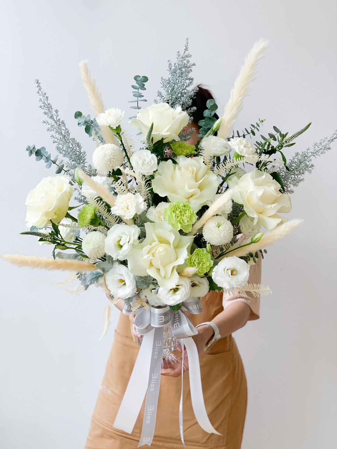 A person in a beige outfit holds a large bouquet of flowers. The bouquet features white and light green roses, fluffy white chrysanthemums, and tall pampas grass, adorned with light green leaves and silver eucalyptus, and is tied with a gray ribbon.