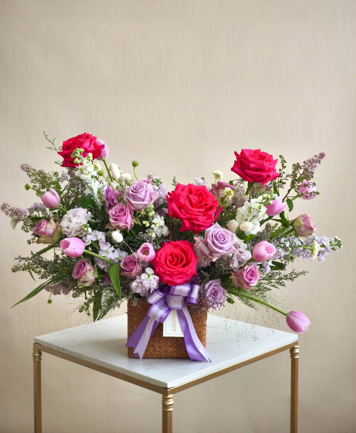 Box arrangement of red roses, purple roses, pink tulips, and mixed seasonal blooms with greenery, tied with a purple ribbon.