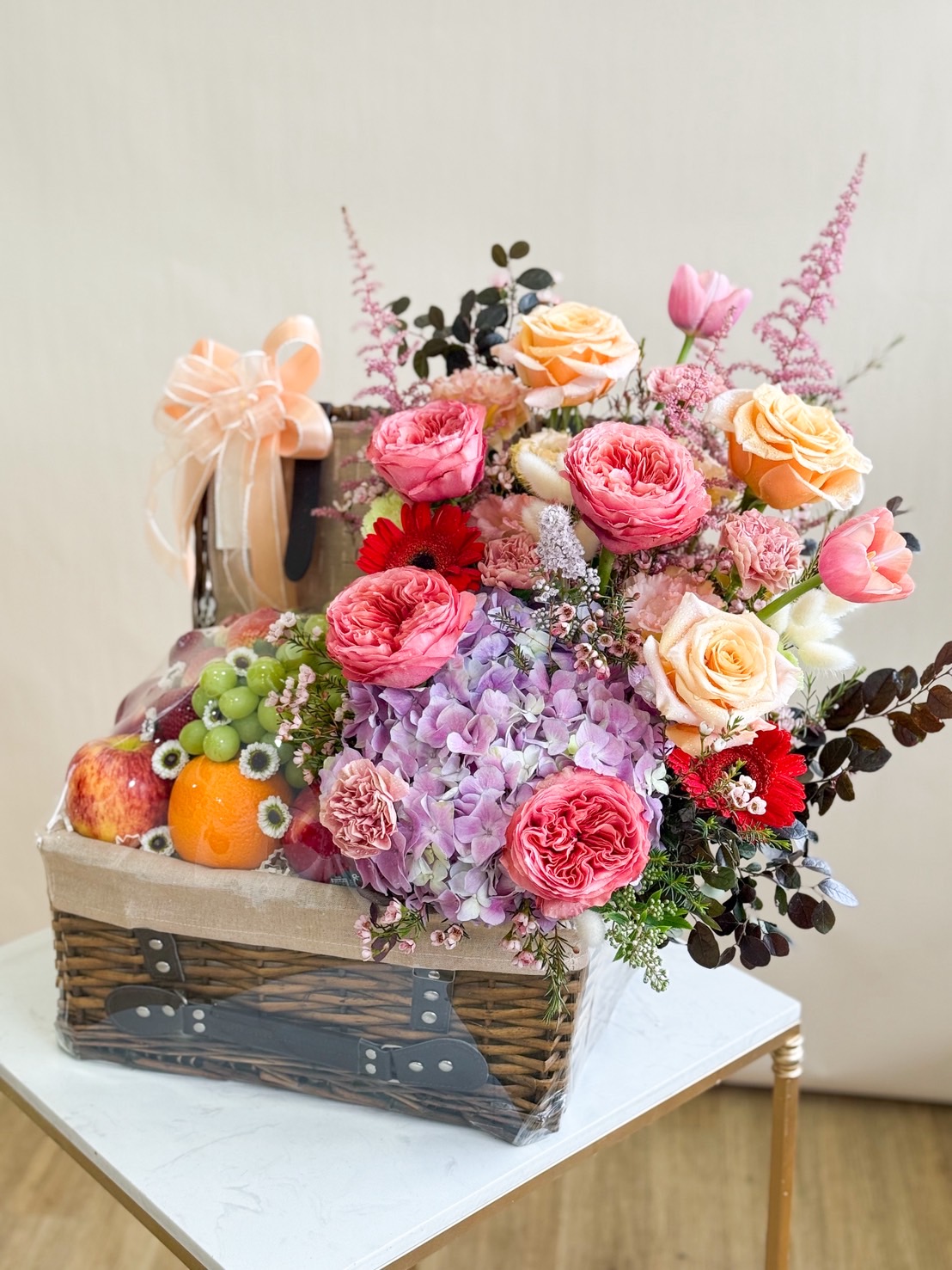 Elegant wicker basket filled with fresh apples, grapes, and oranges, paired with roses, hydrangeas, tulips, and gerberas in warm pink and peach tones.