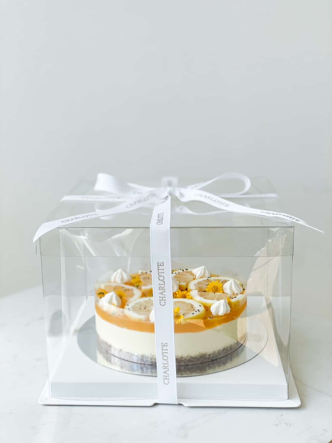 A decorative Passion Fruit Cheese Cake with a white and yellow topping, featuring dollops of cream and edible decorations, is seen inside a transparent box tied with a white ribbon. The cake sits on a white base and is presented on a marble surface.
