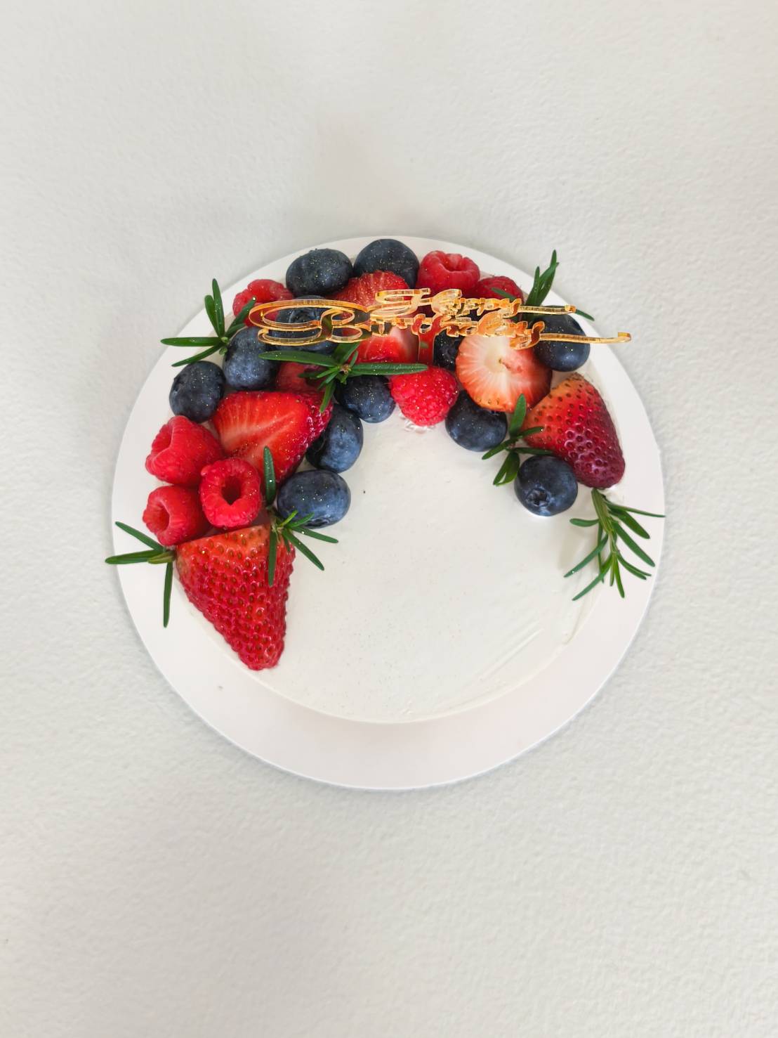 Fresh cream cake topped with strawberries, blueberries, raspberries, and rosemary, decorated with a golden Happy Birthday topper.
