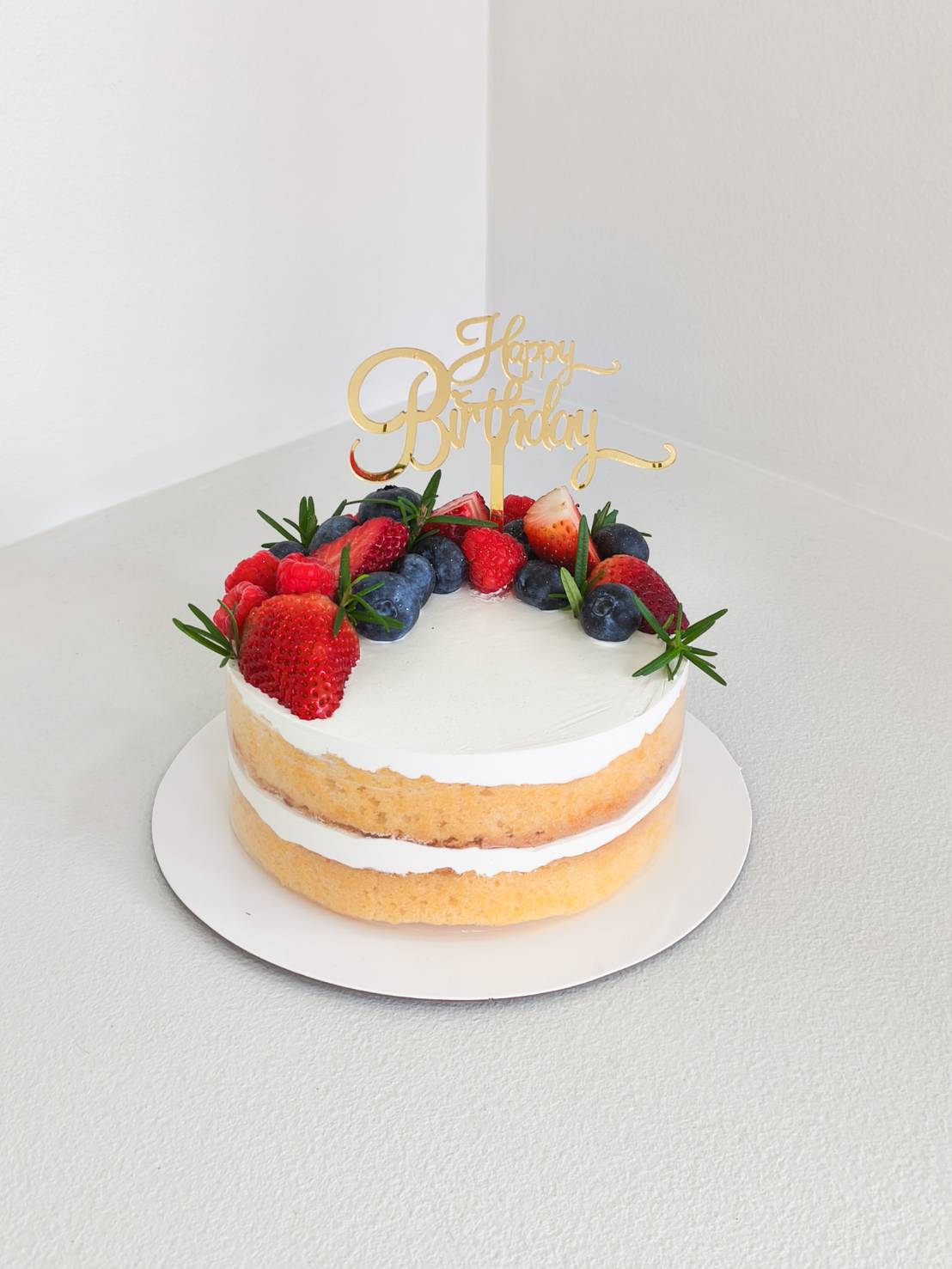 Naked vanilla sponge cake with fresh cream, topped with strawberries, blueberries, raspberries, and rosemary sprigs, with golden Happy Birthday topper.
