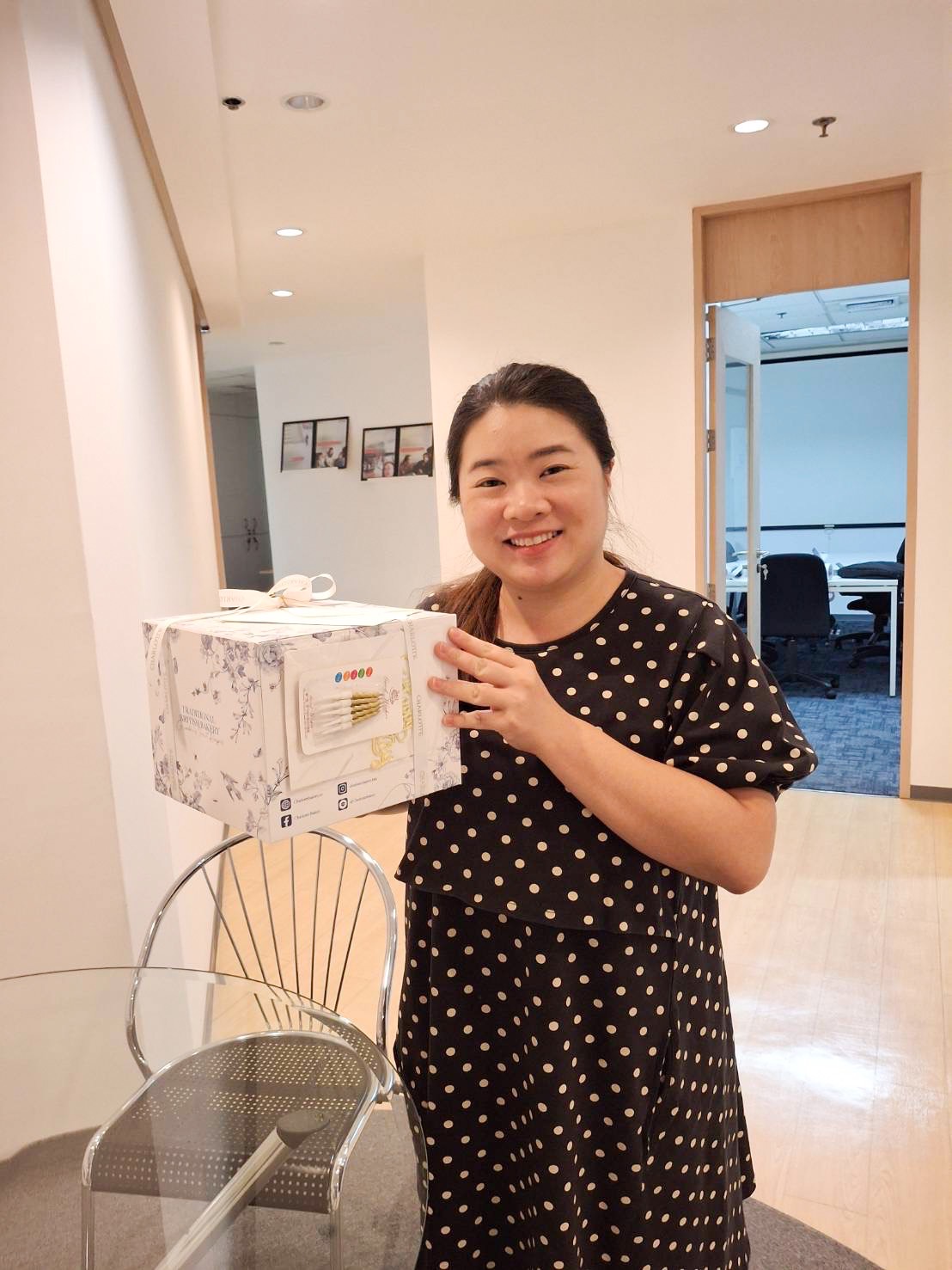 A smiling woman with long dark hair is standing indoors, holding a white gift box wrapped with a floral pattern and a ribbon on top. She is wearing a black dress with white polka dots. Behind her, there's a modern office space with glass walls and framed pictures. She looks excited, perhaps hoping for lemon curd cheesecake inside the box!
