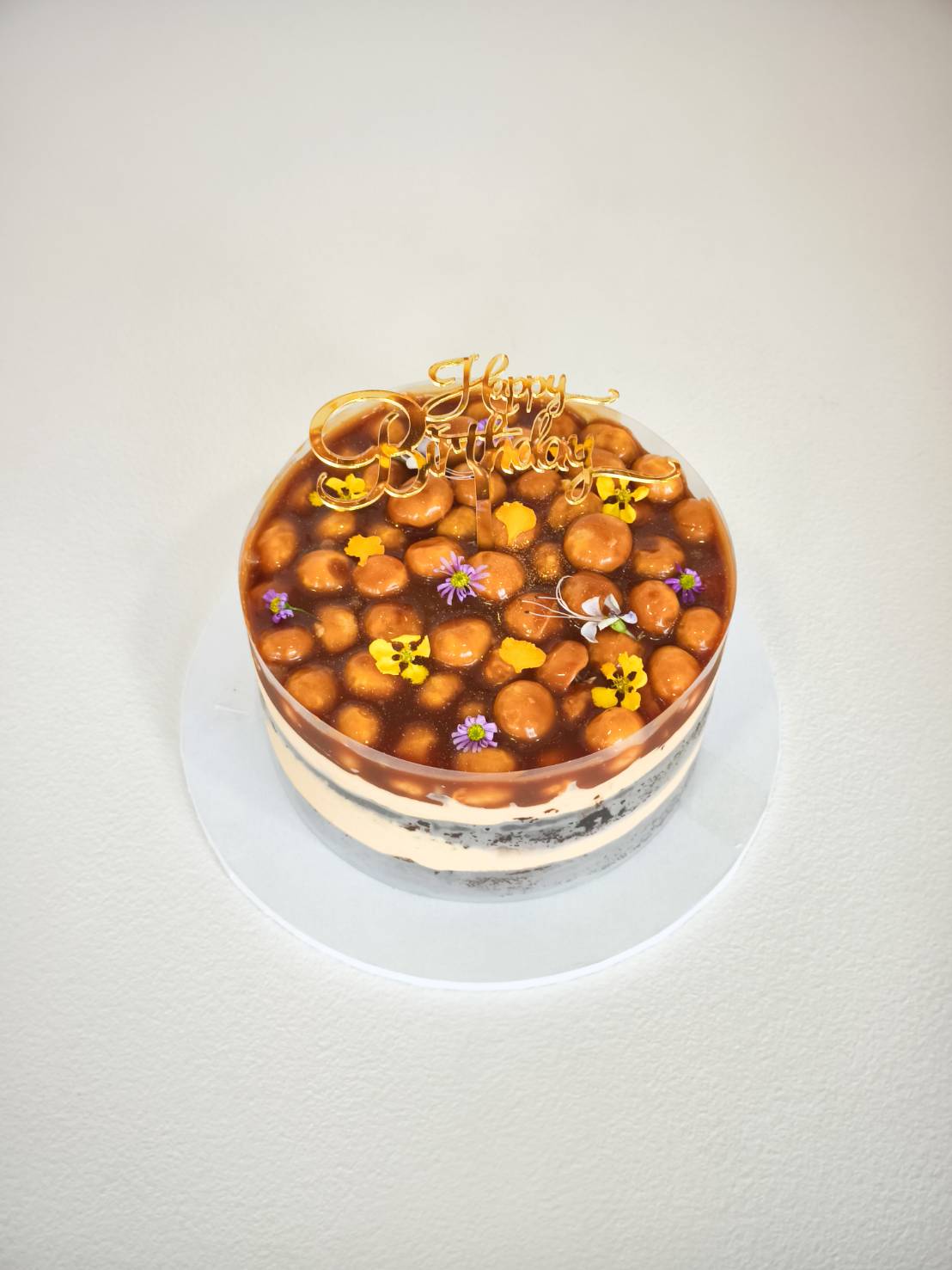 Birthday cake with caramel macadamia nut topping, oreo cream layers, and gold Happy Birthday topper, decorated with edible flowers.