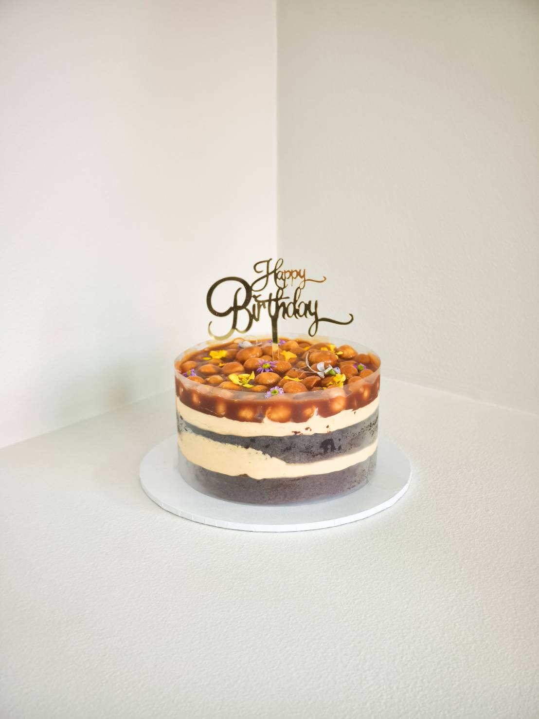 Layered birthday cake with macadamia caramel topping, Oreo base, and vanilla cream, decorated with edible flowers and gold topper.