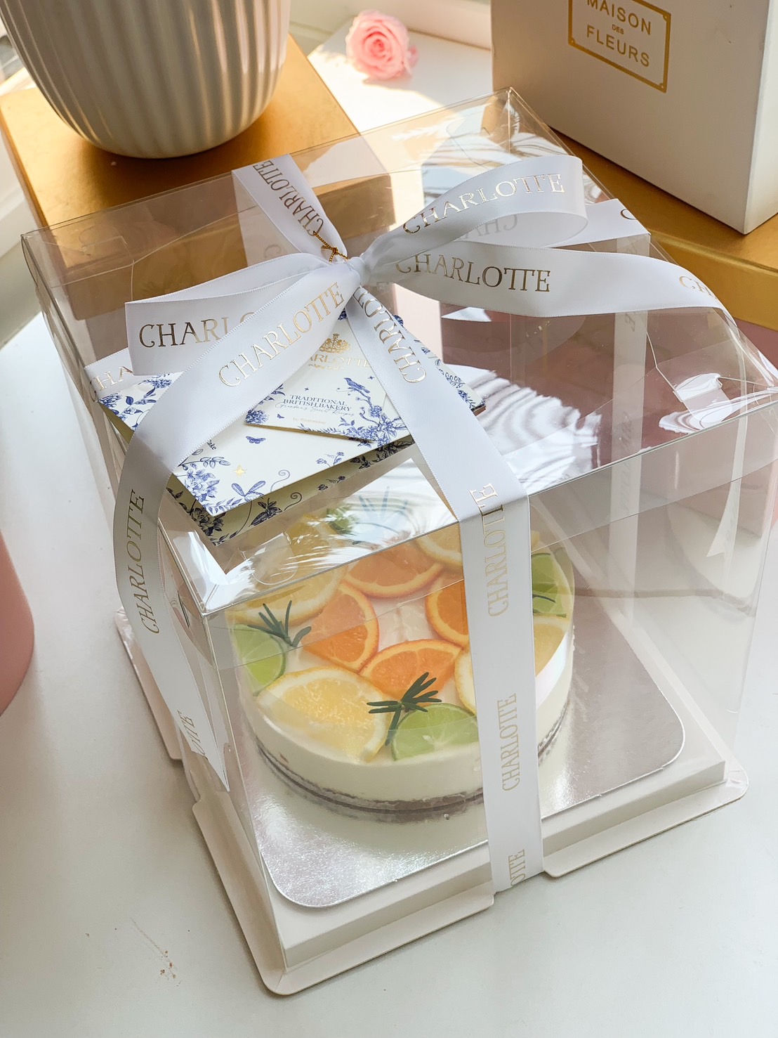 A beautifully packaged citrus cheesecake, topped with fresh orange and lime slices, wrapped with an elegant white ribbon in a clear cake box.