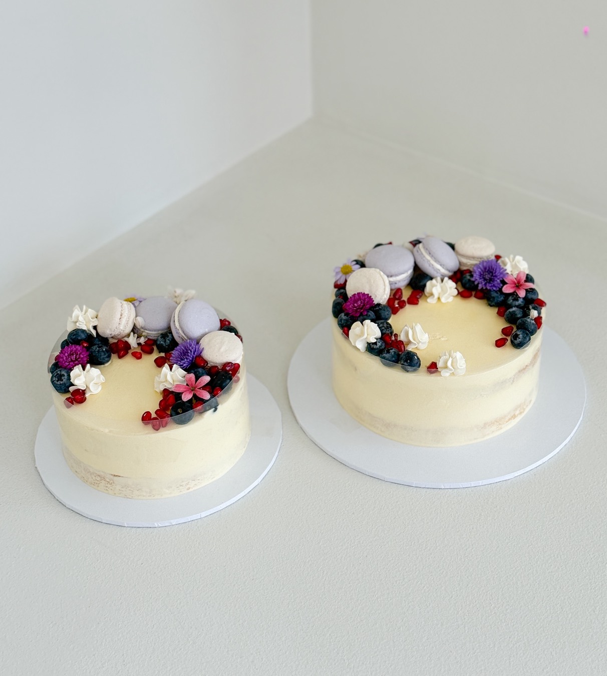 Two buttercream cakes topped with macarons, fresh berries, pomegranate seeds, and edible flowers, placed on white boards.
