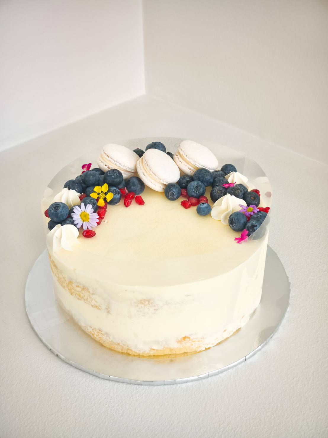 Vanilla cream cake decorated with fresh blueberries, white macarons, edible flowers, and red berries on top.