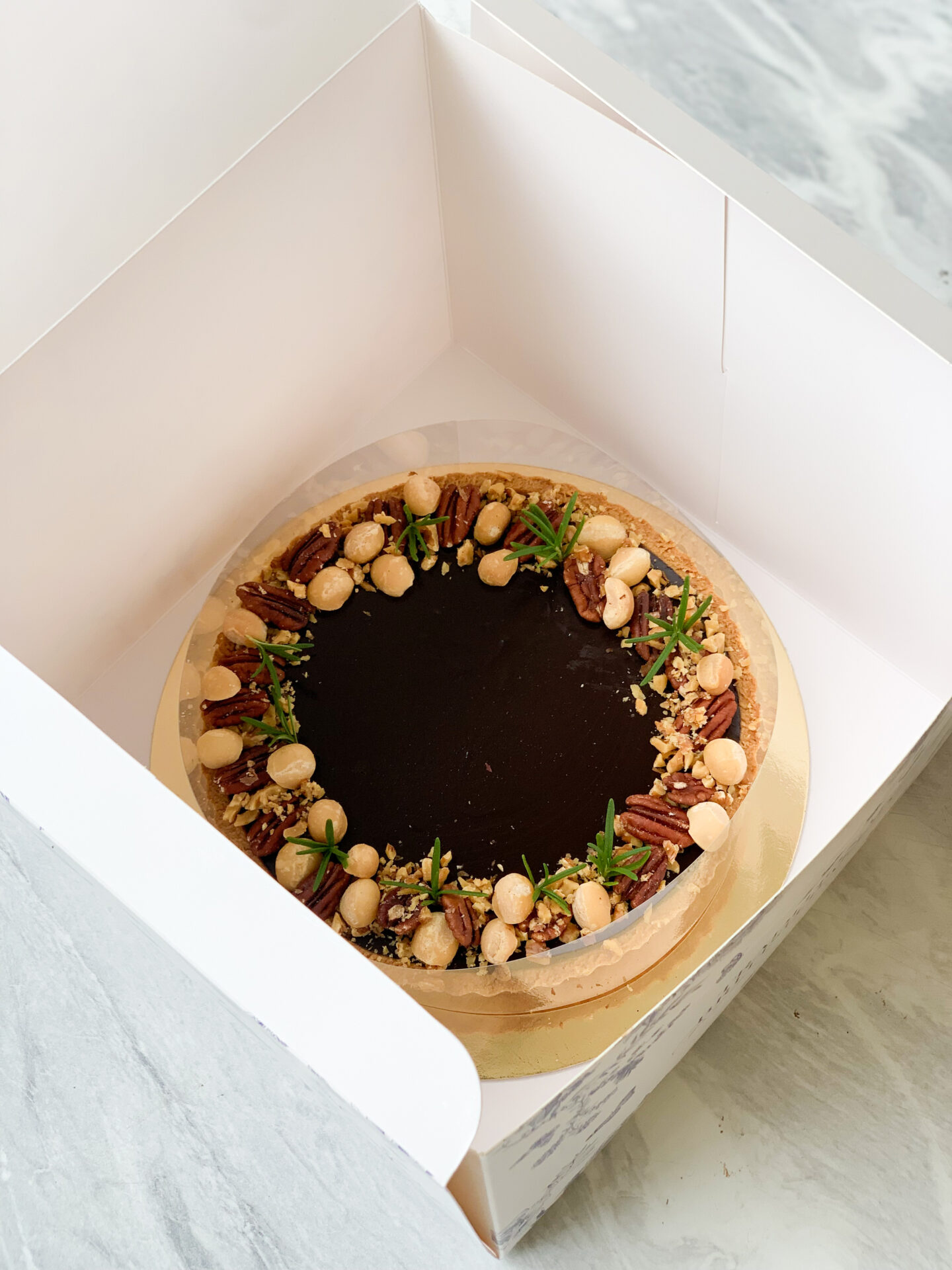 A decadent round dark chocolate tart, adorned with rich macadamias and pecans, rests elegantly within an open white box featuring a sophisticated marble-patterned interior. The glossy surface of the tart glistens invitingly, while delicate green herbs artfully fringe its edge. Displayed prominently is the Basketeer logo on a luxurious ribbon encircling the box, adding an extra touch of opulence to this exquisite dessert presentation.