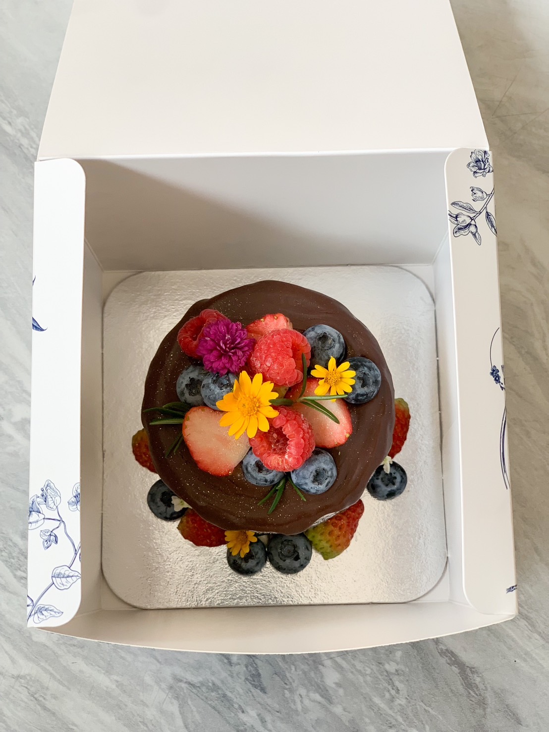 A luxurious chocolate ganache cake topped with fresh strawberries, blueberries, raspberries, and edible flowers, elegantly presented in a premium box.